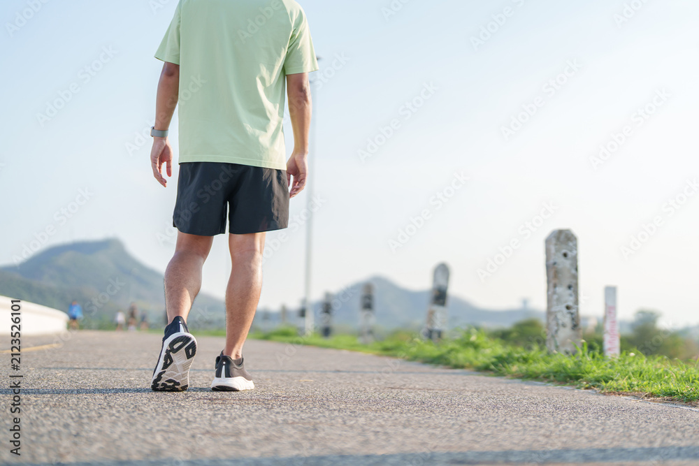 Fototapeta premium Young adult Asian man jogging and walking in the park at morning. male athlete in sport shoes running on the road with visible leg muscles. Healthy lifestyle, wellness and workout concept