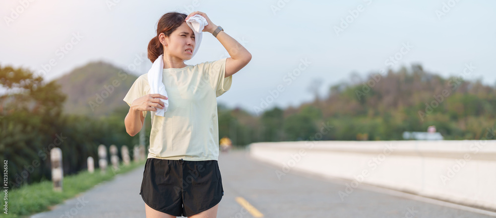 Fototapeta premium Exhausted Asian woman wiping sweat with a towel after jogging in the park. Tired female runner suffering from summer heat and fatigue after fitness workout. Outdoor exercise and heatstroke concept