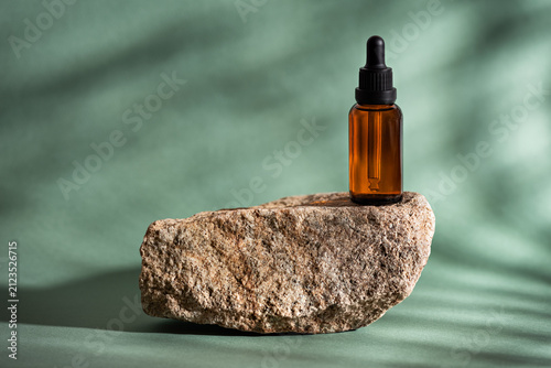 Beauty serum in clear amber glass dropper bottle on stone podium on green background with shadows and light, mockup