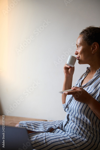 Woman sipping coffee at home in striped dress, relaxed morning portrait with laptop