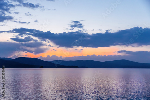 Calm lake reflecting the dramatic sunset with vibrant orange and pink hues beneath layered clouds, silhouetted mountains and a small island in the distance