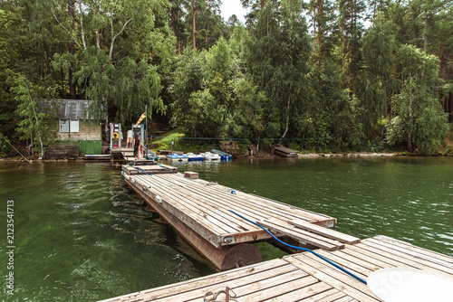 Rustic wooden floating dock extending into calm green water at a forested lakeside recreation area, with a small boathouse, inflatable boats, and people nearby