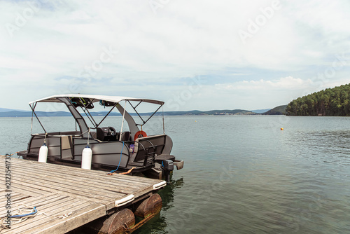 Modern pontoon boat with canopy docked at a weathered wooden pier on a calm lake, with distant forested islands and a cloudy sky, perfect for leisure boating and water recreation.