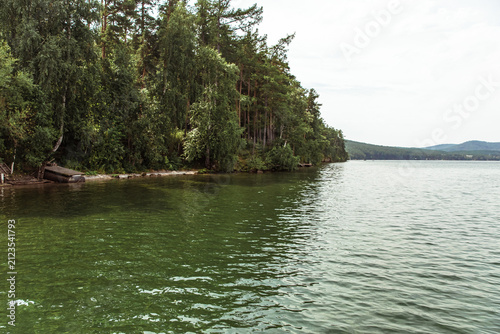 Calm green waters of a large lake bordered by a dense mixed forest of pines and birches, with a small wooden structure on the sandy shore and rolling hills in the distance