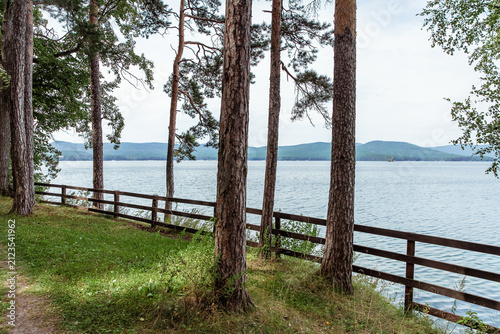 Scenic view of lake framed by tall pine trees and a wooden fence, with rolling hills in the distance and a tranquil water surface