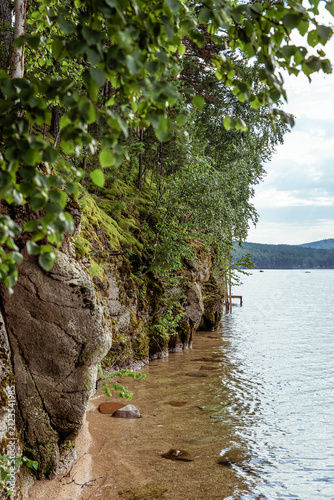 Rocky shore of lake with moss-covered cliffs, green birch foliage, and gentle waves lapping at the sandy beach, showcasing pristine natural scenery.