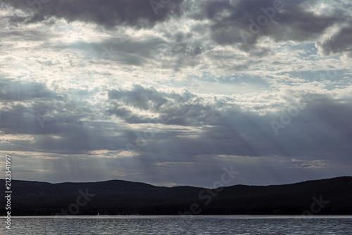 Dramatic cloudy sky with sunbeams over Lake, silhouetted hills in the background and calm water surface, evoking a moody natural atmosphere