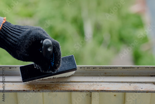 Gloved hand wipes dust and debris from balcony window frame with abrasive sponge before painting, showing careful surface cleaning, outdoor home maintenance, and renovation preparation