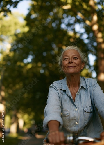 Portrait of an elderly woman riding a bicycle in a park. she is wearing a light blue denim shirt and has short white hair. the woman is smiling and looking up at the sky.