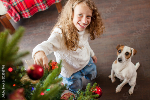 Happy child girl decorating Christmas tree with red ball ornament. Cute dog sitting nearby on floor. Winter holiday celebration at home with pet. Joyful childhood and family festive time.