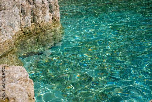 Clear turquoise water gently laps against rocky shore with shimmering sunlight reflections on serene natural coastal landscape.