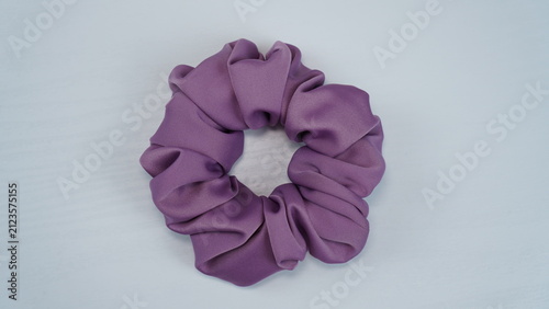 Dusty purple satin scrunchie placed on a white wooden table. Minimal flat lay with soft lighting, elegant hair accessory for fashion, beauty, and lifestyle concepts.