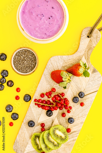 Preparing healthy fruit smoothie. Acai smoothie bowl near cutting board with fresh fruits, berries, chia seeds on yellow background top view