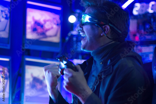 Male gamer, wearing headphones, is playing online video games or competing in esports, displaying a triumphant gesture in a neon-lit gaming room.