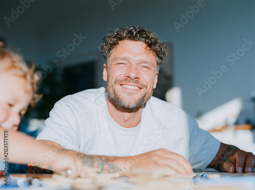 Happy Tattooed Father Laughing with Young Child at Home Dining Table