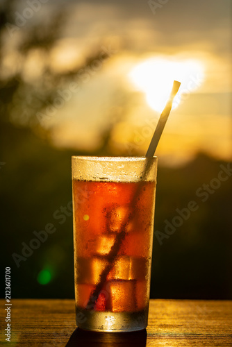 Refreshing iced drink in tall glass with straw against warm sunset background, closeup. Ideal for summer vacation, beach bar menus, cafe advertising, and relaxation concepts