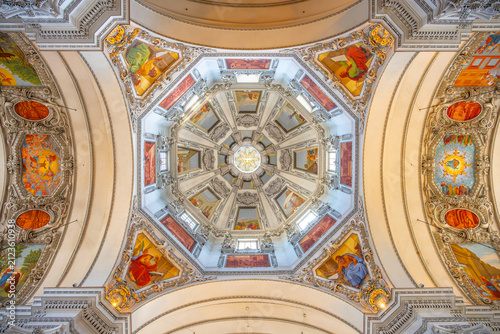 The interior of Basilika St. Lorenz in Kempten. It features a dome with detailed artwork and a bright central light. Visitors admire the architecture and paintings.
