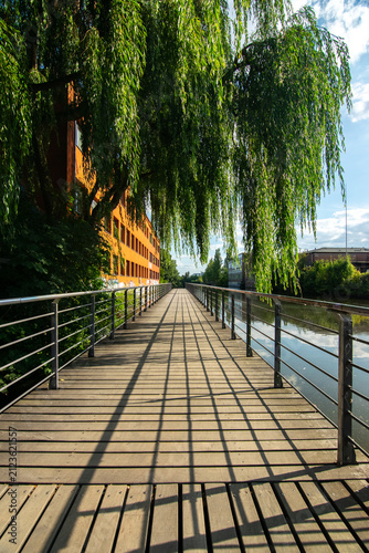 Wooden walkway with shadows next to river and weeping willow