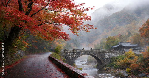 Red autumn maple leaves framing ancient stone arch bridge over misty river in traditional Asian garden