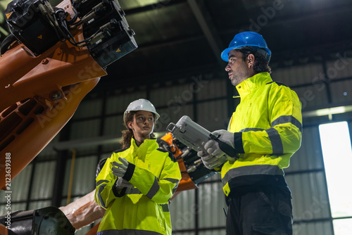 Man and women engineer and worker remote control standing in industrial factory near large manufacturing robotic arm wearing high visibility uniform checking advanced technology during production.