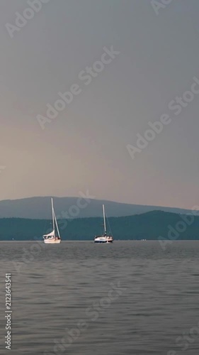 Sailboats anchored on Lake under a moody overcast sky, with distant hills and a subtle sunset glow on the horizon, conveying a tranquil yet dramatic atmosphere