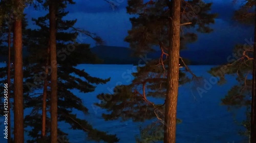 Lightning flashes in the storm clouds, illuminating the mountain lake. Pine trees in the foreground. Blue twilight sky, thunderstorm, twilight