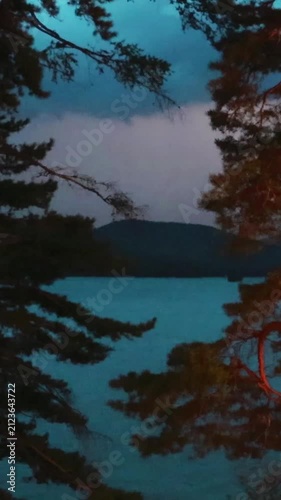 Lightning flashes in the storm clouds, illuminating the mountain lake. Pine trees in the foreground. Blue twilight sky, thunderstorm, twilight
