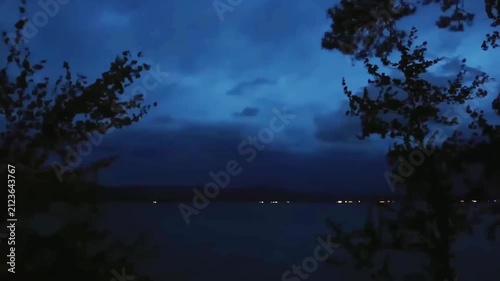 Lightning flashes in the storm clouds, illuminating the mountain lake. In the foreground, trees sway in the wind. Blue twilight sky, thunderstorm, twilight