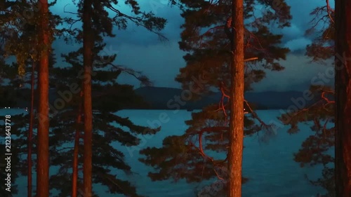 Lightning flashes in the storm clouds, illuminating the mountain lake. Pine trees in the foreground. Blue twilight sky, thunderstorm, twilight