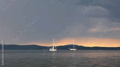 Sailboats anchored on Lake under a moody overcast sky, with distant hills and a subtle sunset glow on the horizon, conveying a tranquil yet dramatic atmosphere