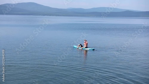 Father and teenage daughter relaxing on a surfboard in shallow water with clear water, smooth rocks visible under the surface. Summer leisure