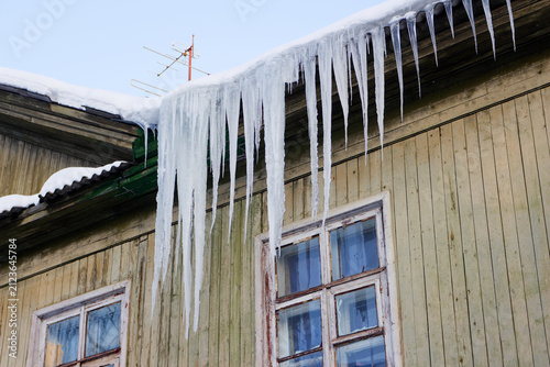 Sharp dangerous icicles hang from the snowy roof edge of an old rustic wooden house during a freezing winter day, creating a beautiful yet menacing frozen natural architecture display