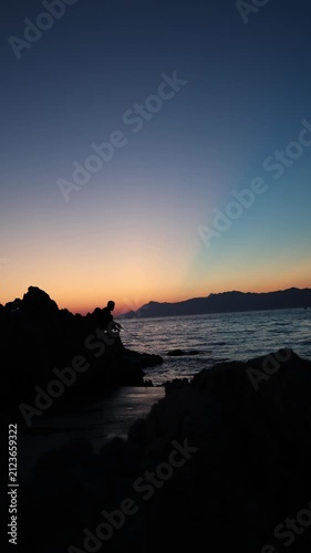 Vertical screen Sunset fishing silhouette on rocky shoreline captures peaceful solitude as an angler waits for the final bite beneath a glowing evening sky by calm sea