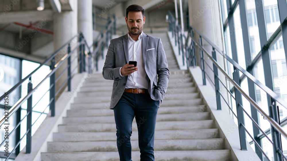 Fototapeta premium Young businessman walking down office building stairs, checking smartphone, staying connected and mobile in his professional life