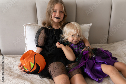 Sisters in witch costumes cuddling on bed holding pumpkin bucket