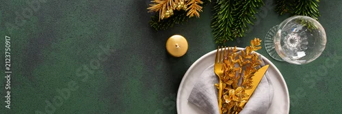 Obraz  z motywem Top view of a Christmas table, featuring an empty plate with a napkin and cutlery on a green surface. Spruse branches and golden tree decorations set a joyful holiday atmosphere, banner