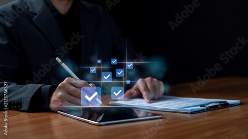 Digital checklist survey and business approval assessment concept. Businessman using stylus pen on tablet with virtual blue checkmark boxes and paperwork documents on desk.