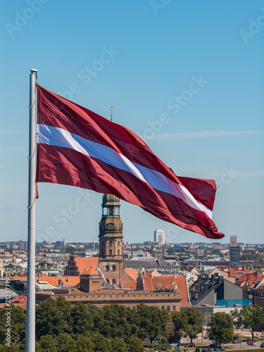 Konstfotografi Aerial drone view of the Latvian national flag flying above the Daugava with the tall green spire of St Peters Church rising from Riga Old Town behind it, under a clear blue summer sky