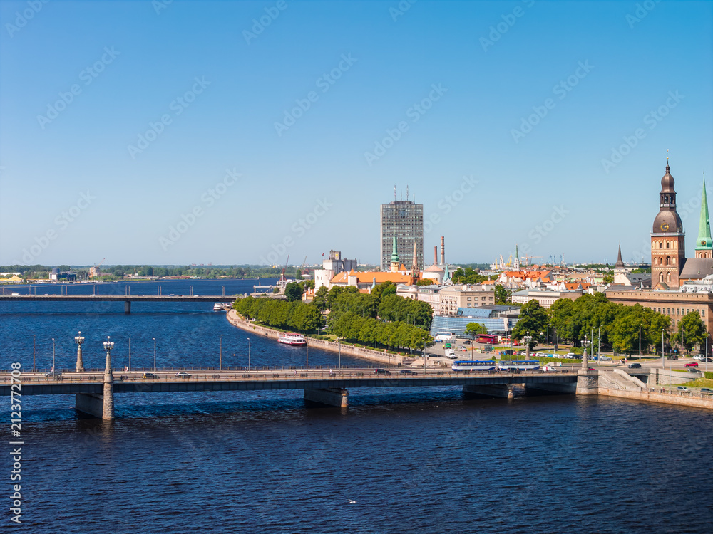 Obraz premium Aerial view of the historic waterfront of Riga Old Town along the Daugava river, with the embankment, tree-lined promenade and the towers of the medieval centre, under a clear blue summer sky.