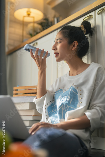 A woman speaks into her smartphone using a voice command feature, sitting comfortably in a warm, softly lit kitchen nook surrounded by books and cozy home decor, blending everyday life with smart tech