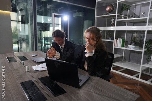 Принти на полотні Nervous business duo wait for critical results at a laptop late at night, the man clasping his hands at his lips and the woman touching her collar anxiously in a bright modern coworking