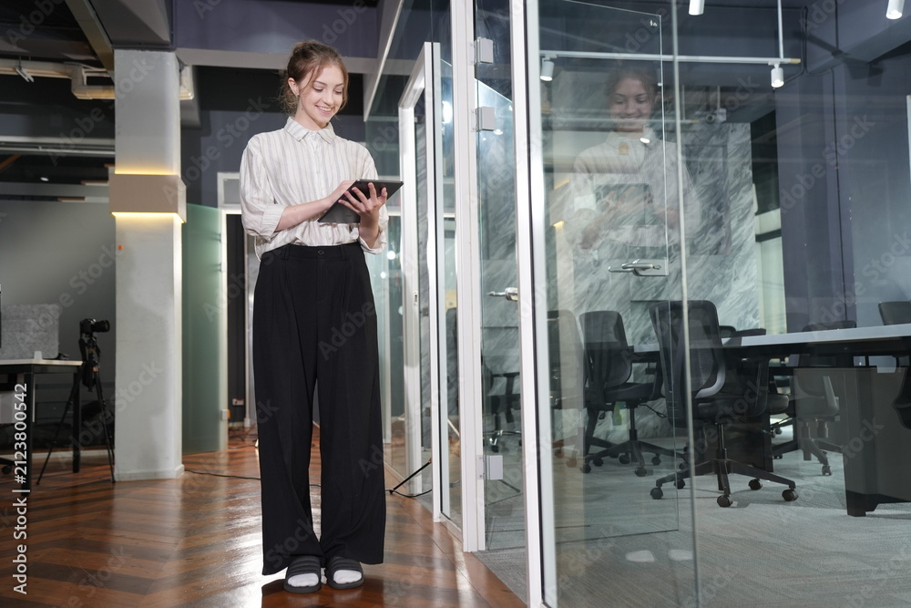 Fototapeta premium Young businesswoman holds a tablet close and smiles gently while standing in a glowing glass-lined corridor of a dim modern coworking office late in the evening.