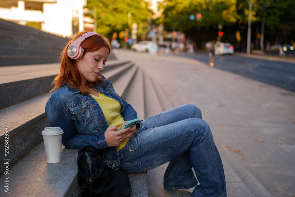 Fototapeta premium Young woman relaxing on city steps, listening to music with pink headphones and using smartphone