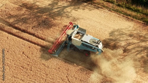 Farmer harvesting ripe barley with combine harvester across large cereal field. Grain filling hopper while straw spreading behind working machine. Modern agriculture farm equipment and rural food