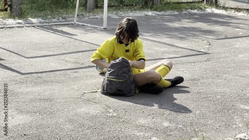 Teen boy sitting on outdoor court with backpack searching inside after football training rest moment youth sport lifestyle and routine concept. High quality FullHD footage