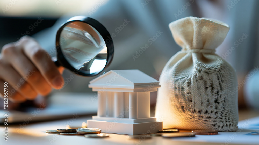 Obraz premium Close-up of hand holding magnifying glass over bank model and money bag, financial investigation and anti-money laundering compliance concept, defocused background, with copy space