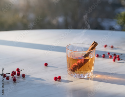 Steaming hot festive winter drink garnished with cinnamon stick, orange peel, and red berries on a bright sunlit marble table