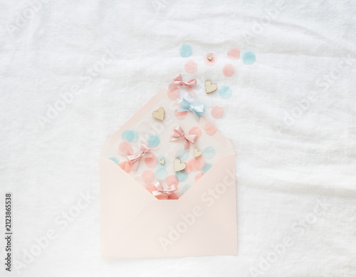 Light pink envelope gently reveals a charming assortment of pastel confetti, delicate bows, and small wooden hearts scattered on a soft white textile background