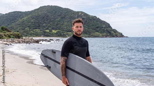 Portrait of a handsome young man holding a surfboard on a beach with a mountain in the background