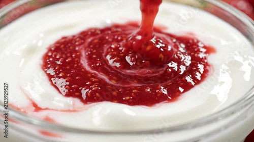 Raspberry jam being poured into creamy yogurt in a glass bowl, close-up slow motion